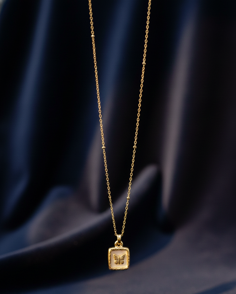 Gold necklace with a square pendant featuring a butterfly design on a dark blue fabric background.